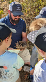 An adult man shows four Tiger Scouts an abandoned bird's nest outdoors